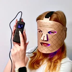 Osoba w beżowej masce LED do fototerapii trzyma kontroler urządzenia, maska świeci na fioletowo pod oczami.