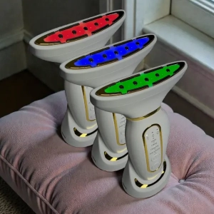 Trzy urządzenia beauty do pielęgnacji twarzy z terapią LED – czerwone, niebieskie i zielone światło – ustawione obok siebie na poduszce przy oknie, nowoczesny design w białej obudowie ze złotymi detalami.